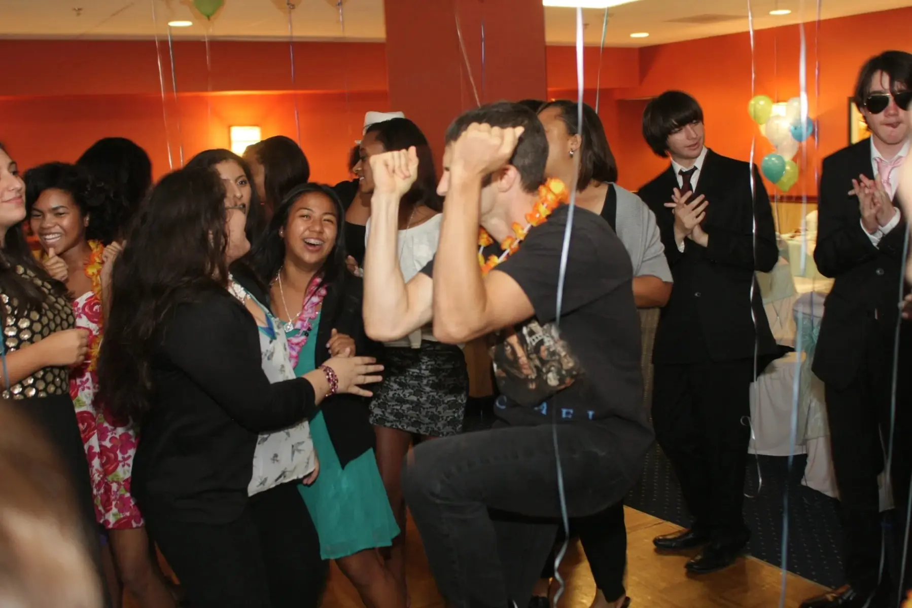 s2 A person dances energetically in a room decorated with balloons, surrounded by smiling people, some in formal attire.