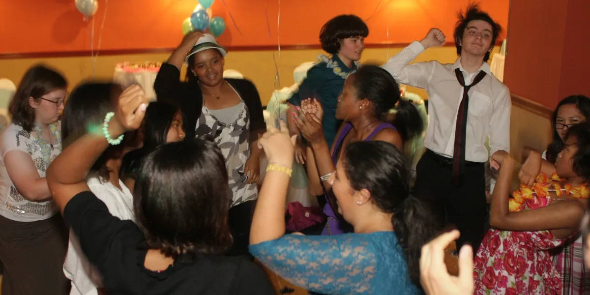 rs=w_1200,h_600,cg_true (1) A group of teenagers dances and socializes in a decorated room with balloons and orange walls.
