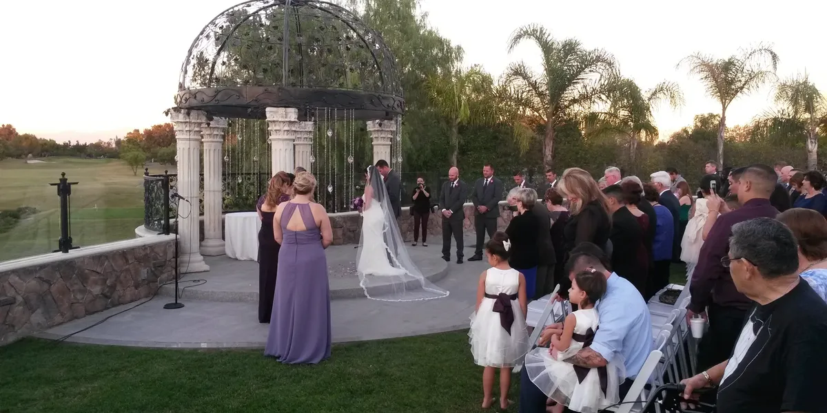 rs=w_1200,h_600,cg_true A wedding ceremony outdoors with a couple at the altar under a gazebo, guests seated and standing, and flower girls in white dresses in the foreground.