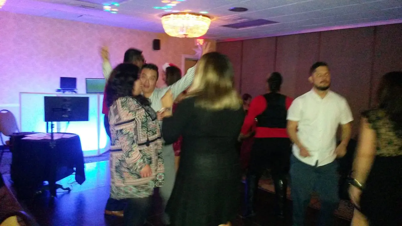 Circa - 2018 Christmas Holiday Party A group of people are dancing and socializing in a dimly lit room with a chandelier and a DJ setup in the background.