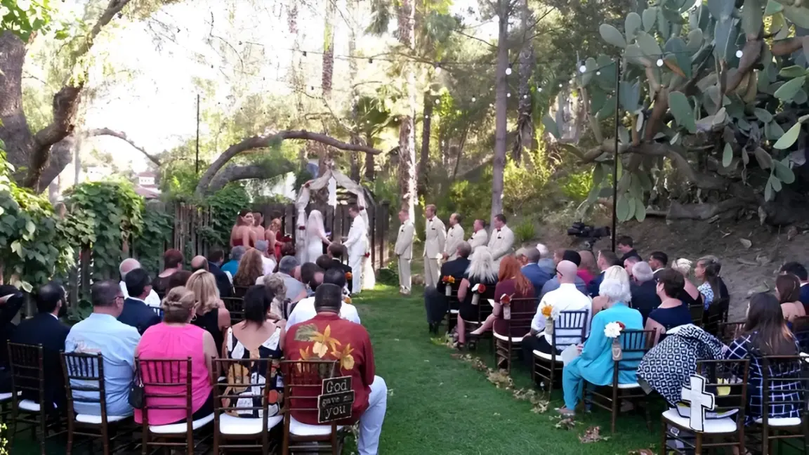 rs=w_1280 (2) Outdoor wedding ceremony with a bride and groom under an arch, surrounded by a wedding party in matching attire. Guests are seated on either side in a garden setting with trees and lights.