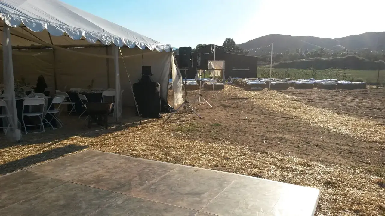 rs=w_1280 Outdoor event setup with a white tent containing chairs and tables on the left, a speaker and bales of hay on the right, and a wooden floor in the foreground, under a clear sky.