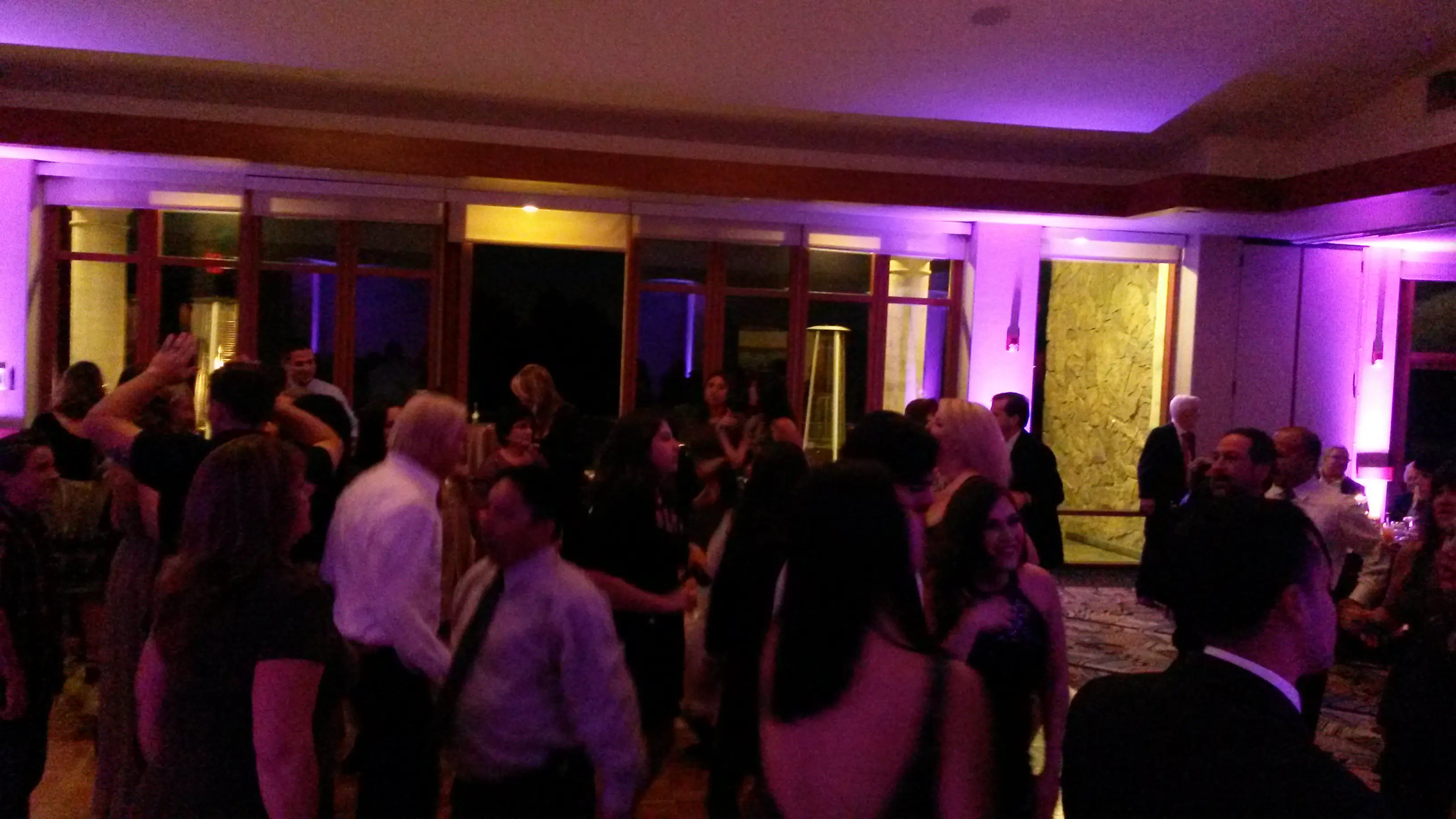 Mr Dj Services Wedding Dj A group of people are dancing and socializing in a dimly lit room with purple lighting and large windows in the background.
