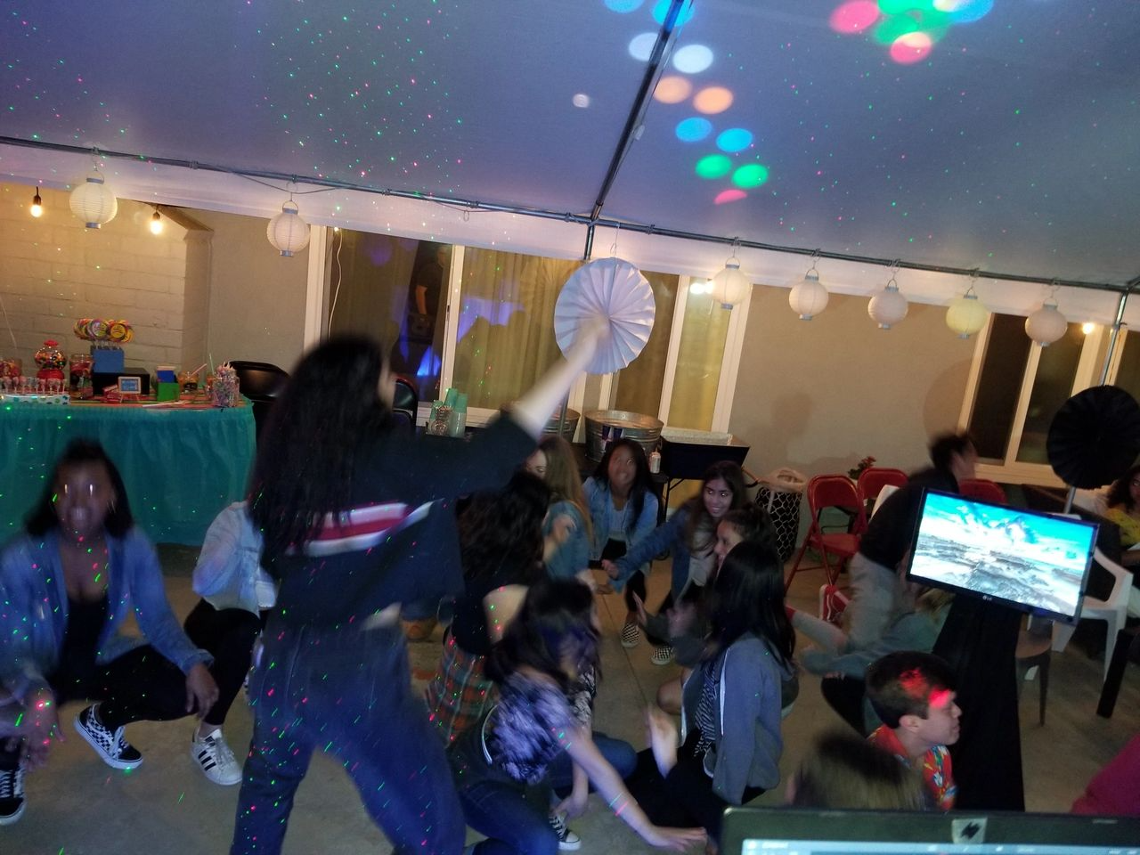 rsw1280 (1) A group of people, mostly children, dance under a tent with string lights and colorful disco lights at a party. A decorated table and large paper fans are visible in the background.