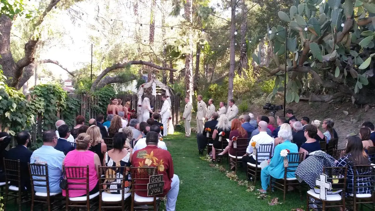 rs=w_1280 (1) Outdoor wedding ceremony with a bride and groom at the altar, bridesmaids and groomsmen standing by, and guests seated on either side of the aisle in a garden setting.