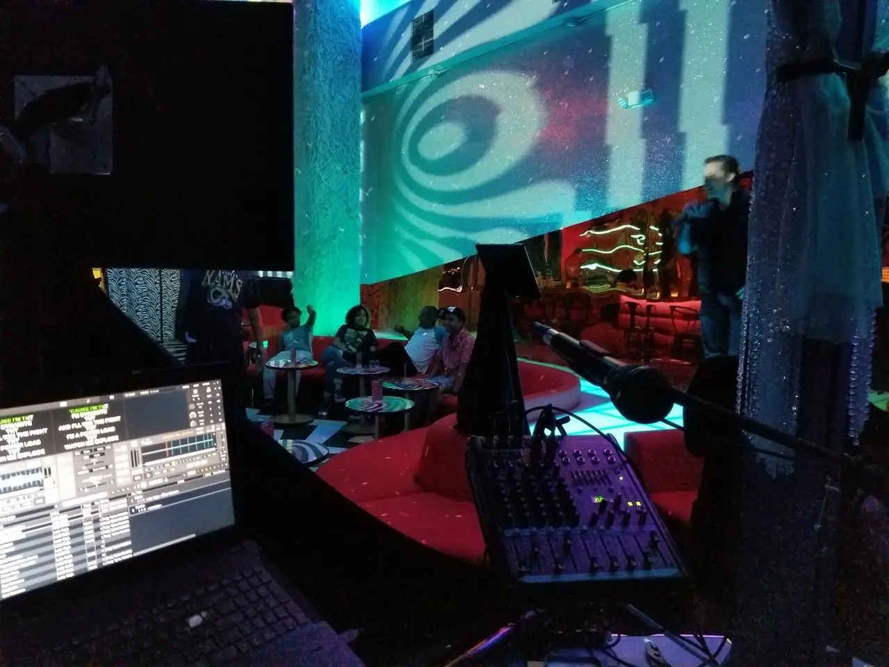 Mr Dj Services - Karaoke Dj A DJ booth with a sound mixer and laptop overlooks a lounge where people are seated on red sofas while a person speaks or sings onstage under colorful lights.