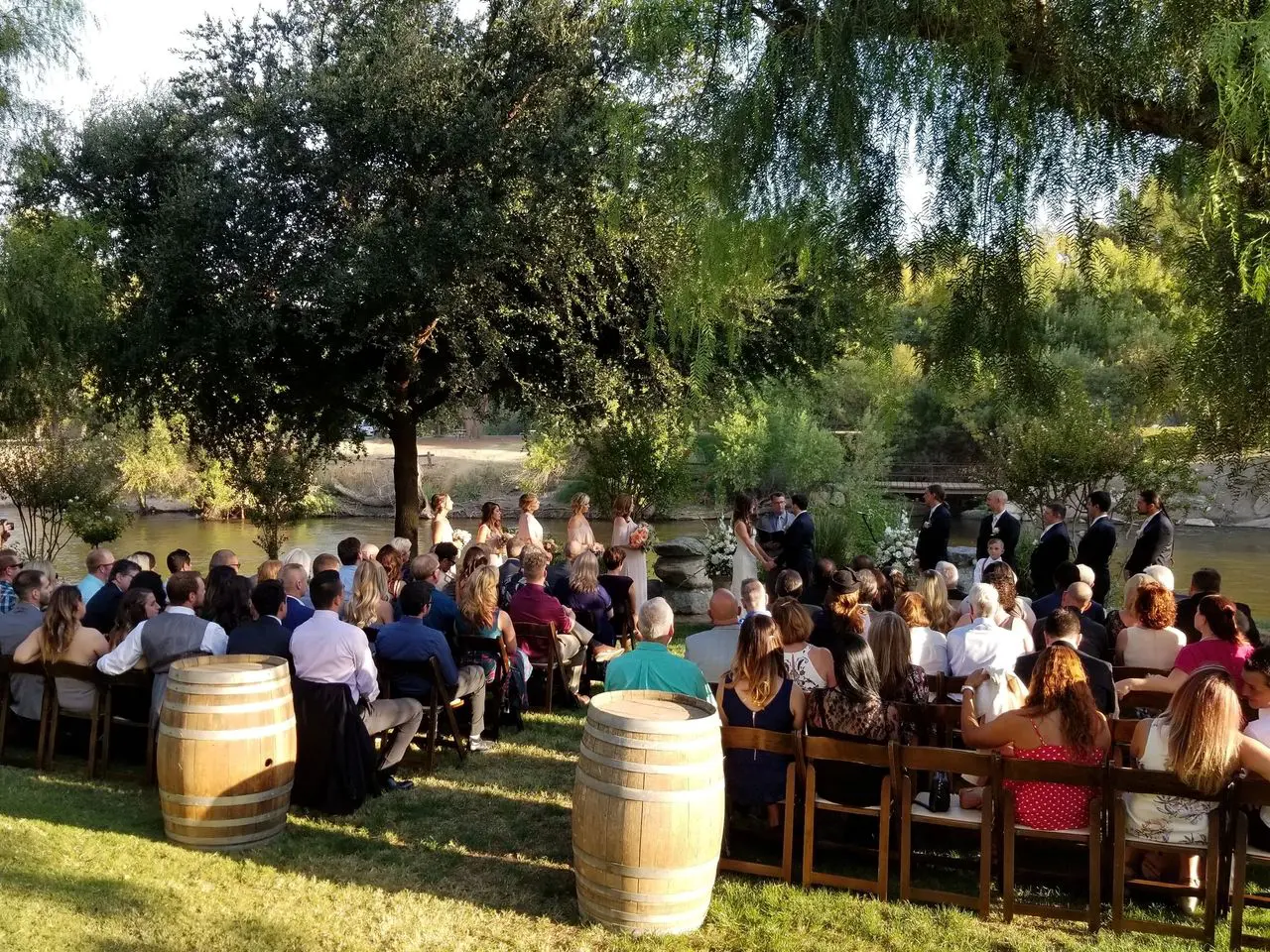 rs=w_1280 (10) Outdoor wedding ceremony by a river with guests seated on wooden chairs, a wedding party at the front, and wine barrels in the foreground under trees.