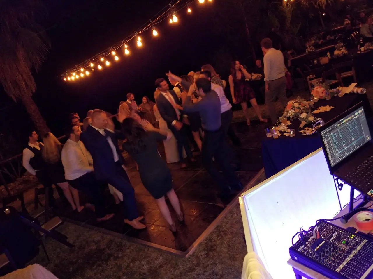 best wedding dj | los angeles wedding djs A group of people dance on an outdoor dance floor at night under string lights, with a DJ setup and sound equipment visible in the foreground.