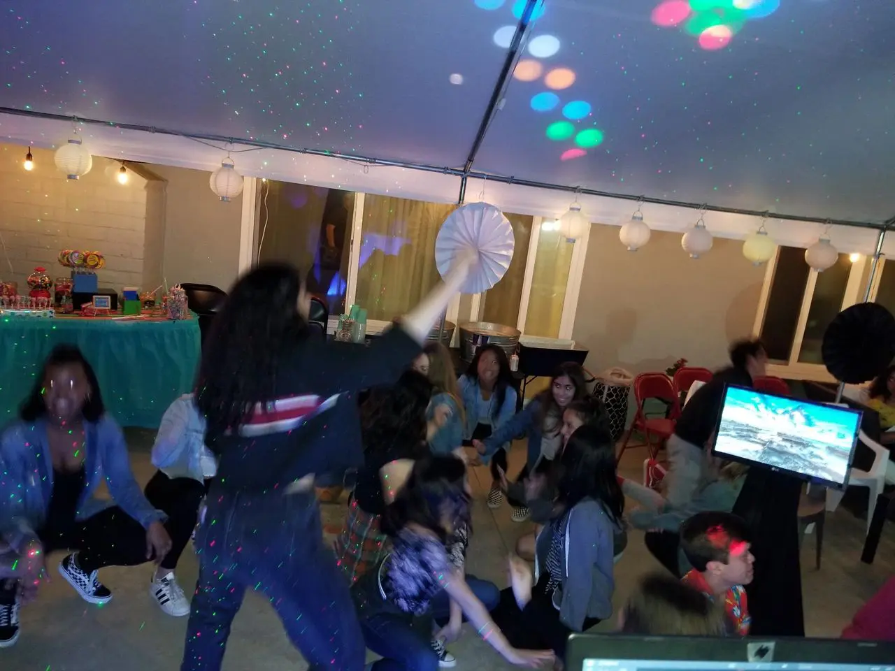 Mr Dj Services kids karaoke A group of people dance under colorful lights at an indoor party, with decorations and a table of snacks in the background.