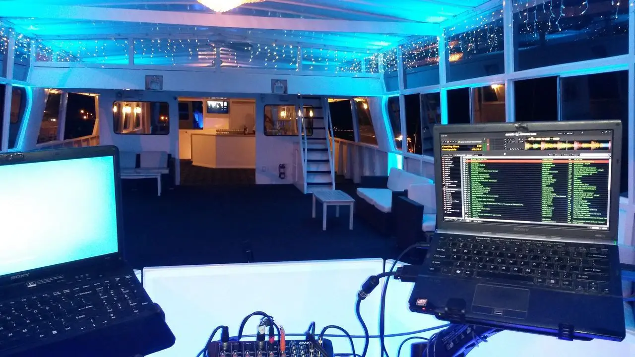 karaoke dj party birthday A DJ setup with two laptops and a mixer sits at the front of a brightly lit event space on a boat, featuring blue lighting, benches, and string lights on the ceiling.