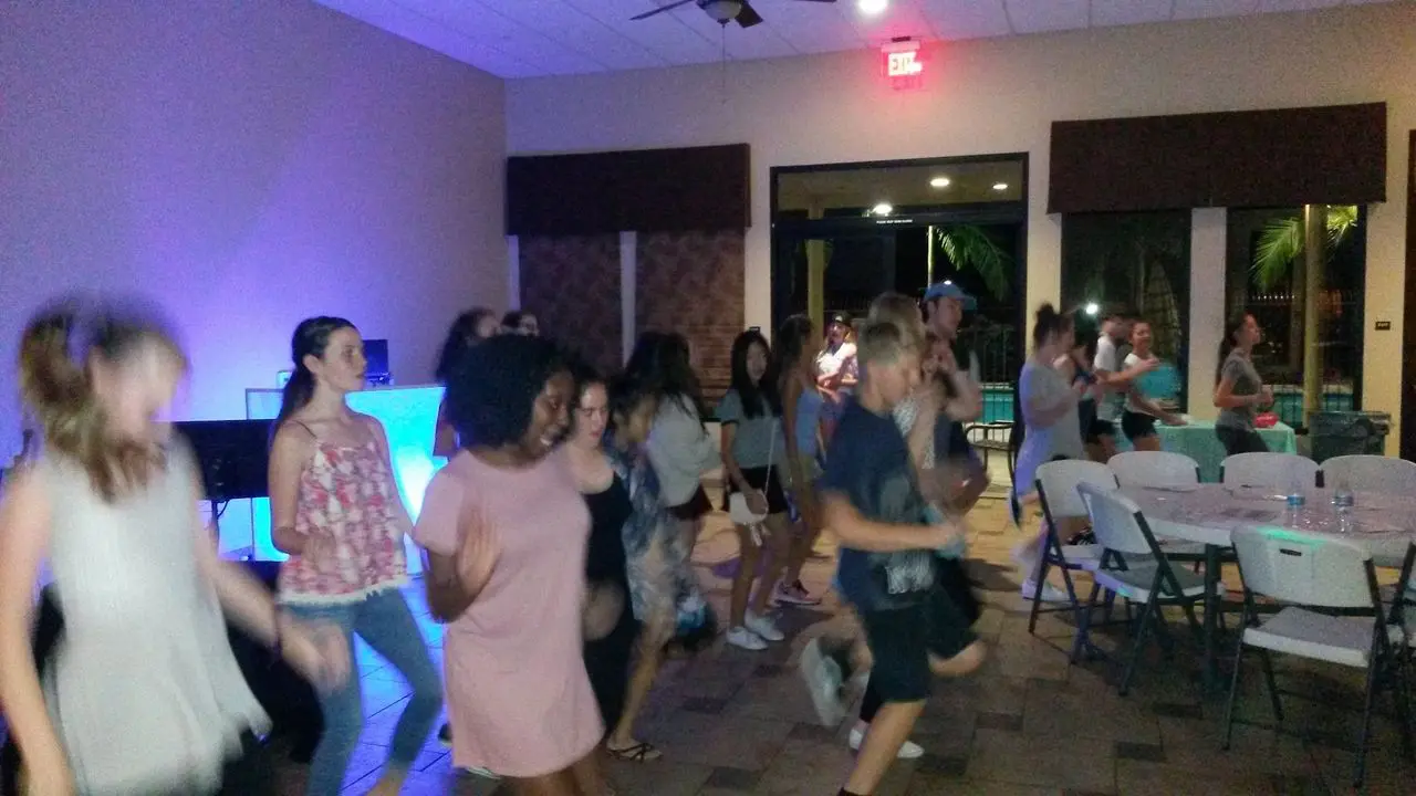 Karaoke Dj Party A group of people are dancing indoors in a well-lit room with tables and chairs set up around them.