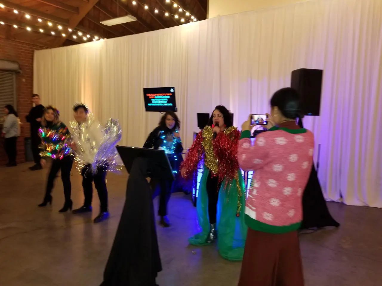 karaoke dj party Four people perform karaoke in festive, shiny costumes while another person in a sweater films them with a phone in a room decorated with string lights and white curtains.