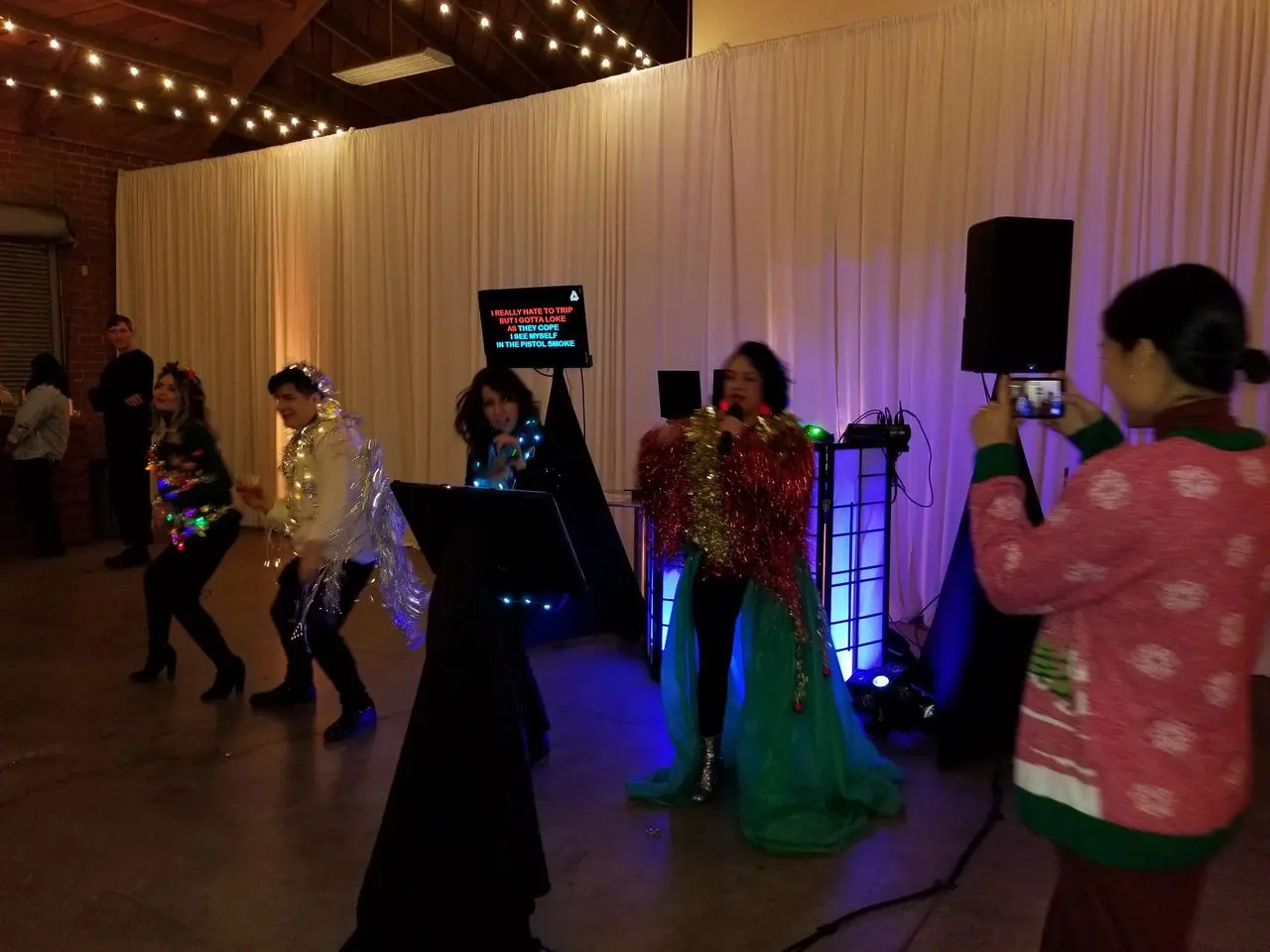 Ugly Sweater Karaoke Dj Party Four people in colorful, shiny costumes perform karaoke on a stage, while a woman in a festive sweater takes a photo with her phone. White drapes and string lights decorate the venue.