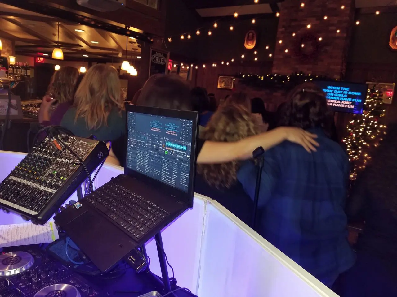 karaoke dj A group of people with arms around each other sing karaoke at a bar, viewed from behind a DJ booth with audio equipment and a laptop.