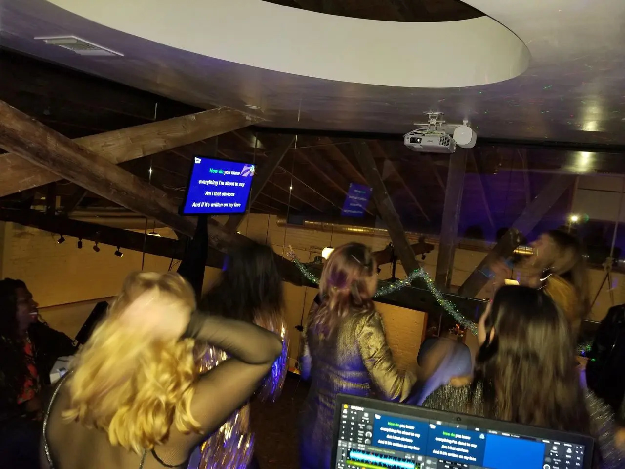 karaoke dj party A group of people sing karaoke in a dimly lit room, facing a screen with song lyrics projected, under exposed wooden beams and a circular ceiling feature.