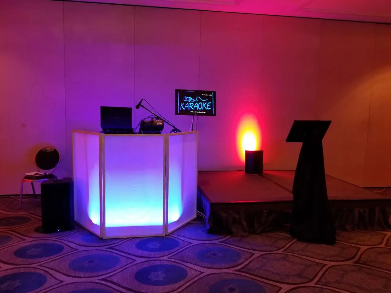 Unknown Karaoke Dj Location A karaoke setup with a lit DJ booth, microphone, laptop, monitor displaying “KARAOKE”, and a small stage with a music stand in a room with patterned carpet.