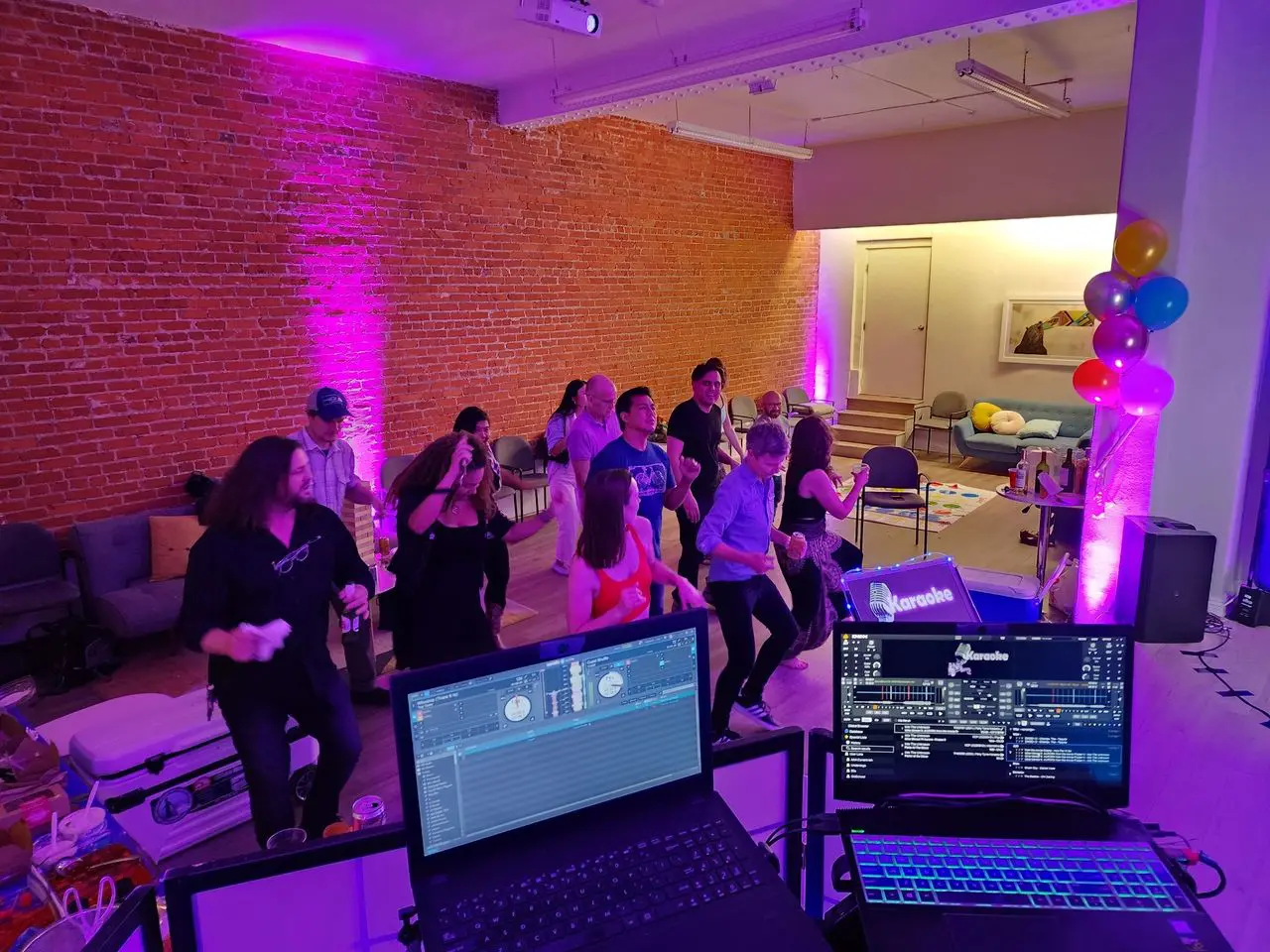 Mr Dj Services A group of people dance in a room with exposed brick walls and purple lighting, viewed from behind DJ equipment. Balloons and snacks are visible in the background.