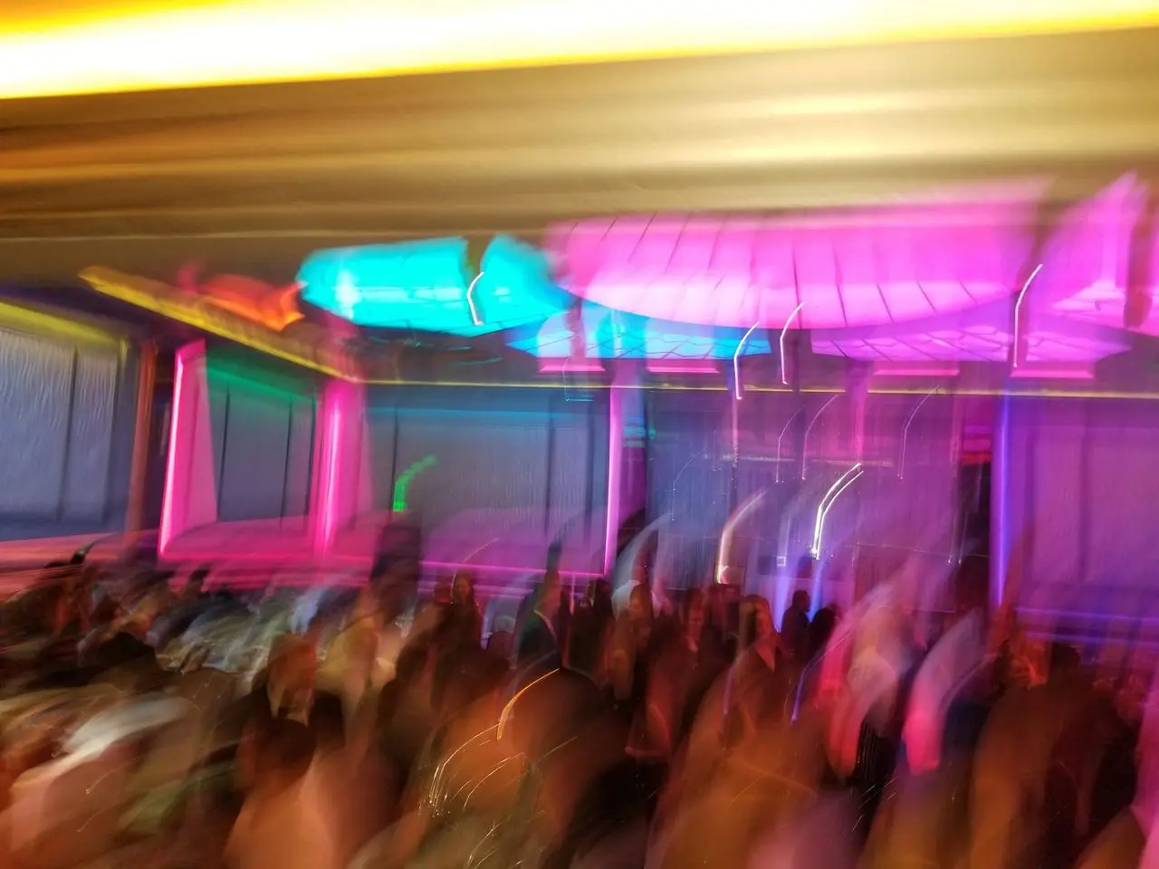 Mr Dj Services - Spanish Dj A blurred image shows a crowd under vibrant neon lights in shades of pink, blue, and yellow, creating an abstract effect.