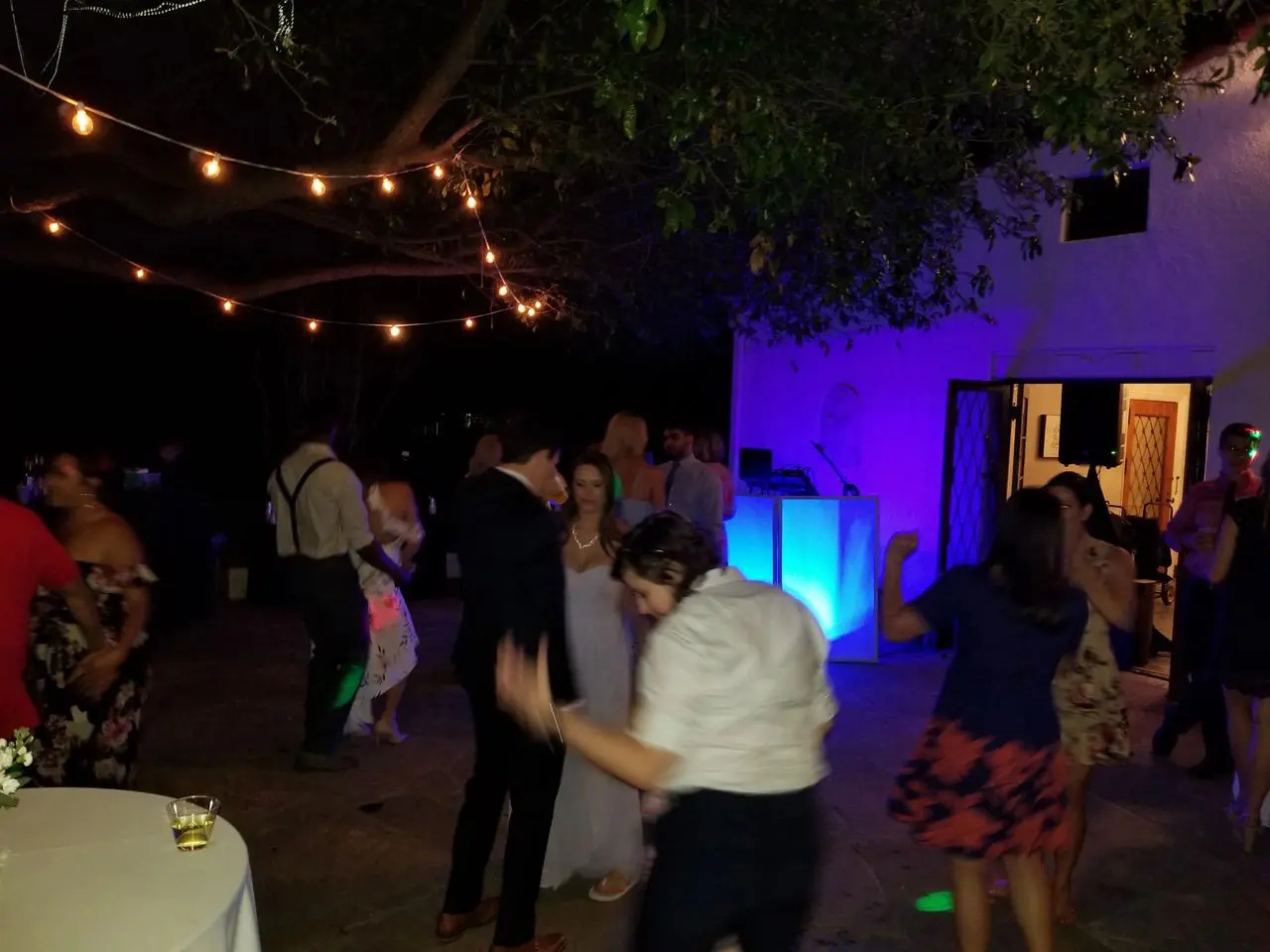 dj services, los angeles dj services People dance outdoors at night under string lights, with a lit DJ booth and a white building in the background. Drinks and tables are visible in the scene.