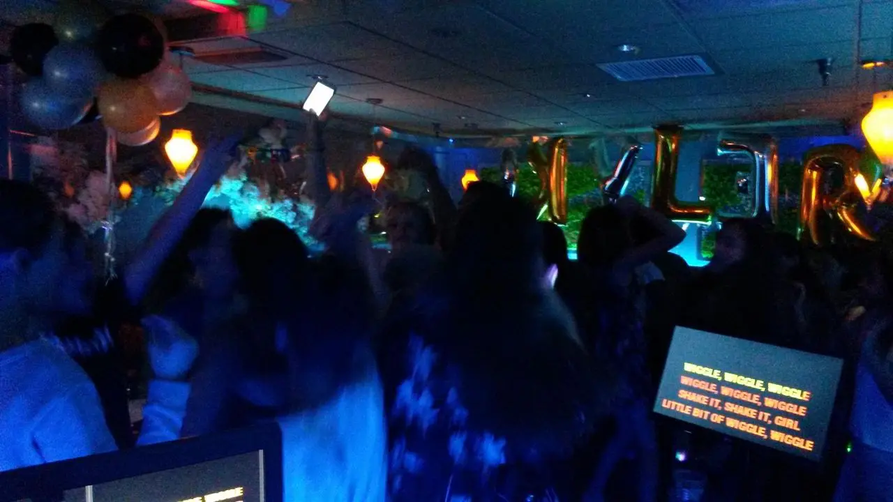 karaoke dj, los angeles dj A group of people dance in a dimly lit room decorated with balloons and gold letter balloons, while karaoke lyrics are displayed on a screen in the foreground.