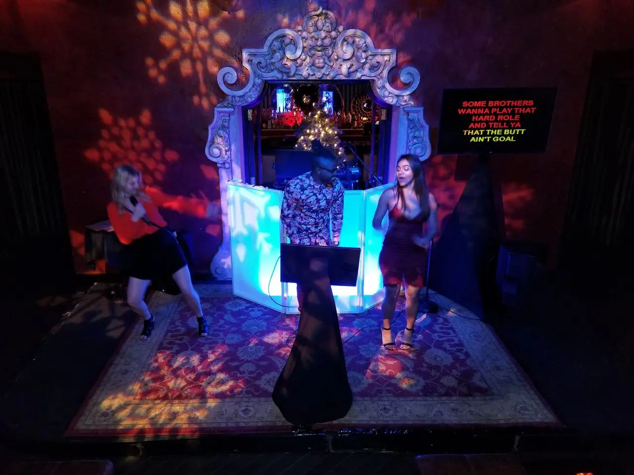 Actor Coach Killian Three people are performing on a small stage with a decorative backdrop and colorful lights; lyrics are displayed on a screen to the right.