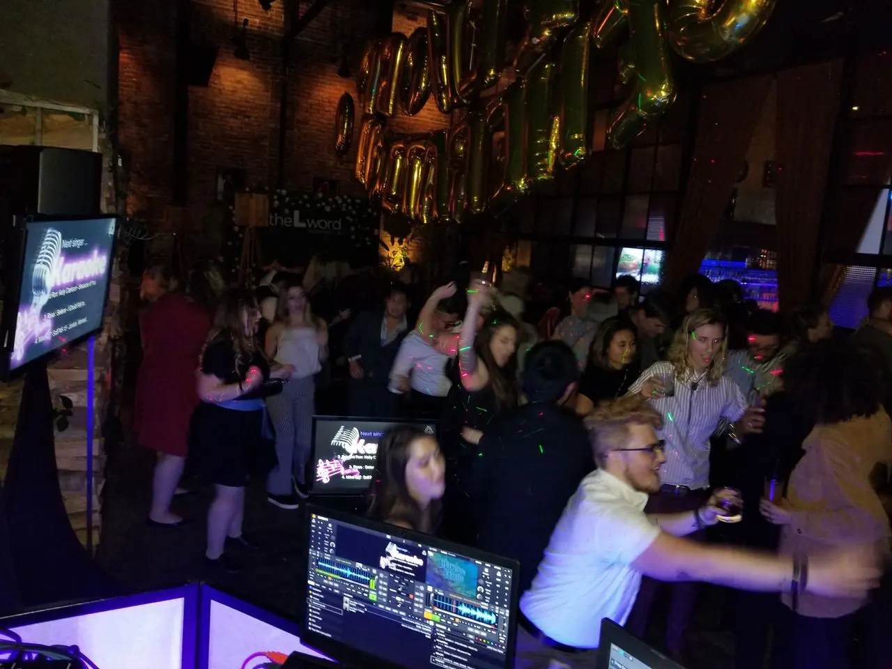 Mr Dj Services - Karaoke dj A crowd of people dance and socialize at a dimly lit indoor party with gold balloons and a karaoke setup visible in the foreground.