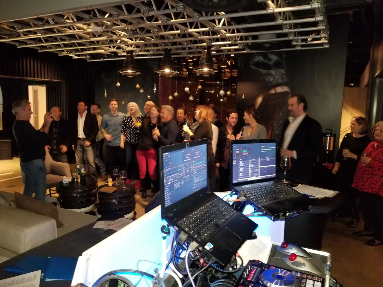Corporate Karaoke Dj party A group of people in semi-formal attire gather and socialize at an indoor event space, with DJ equipment and laptops set up in the foreground.