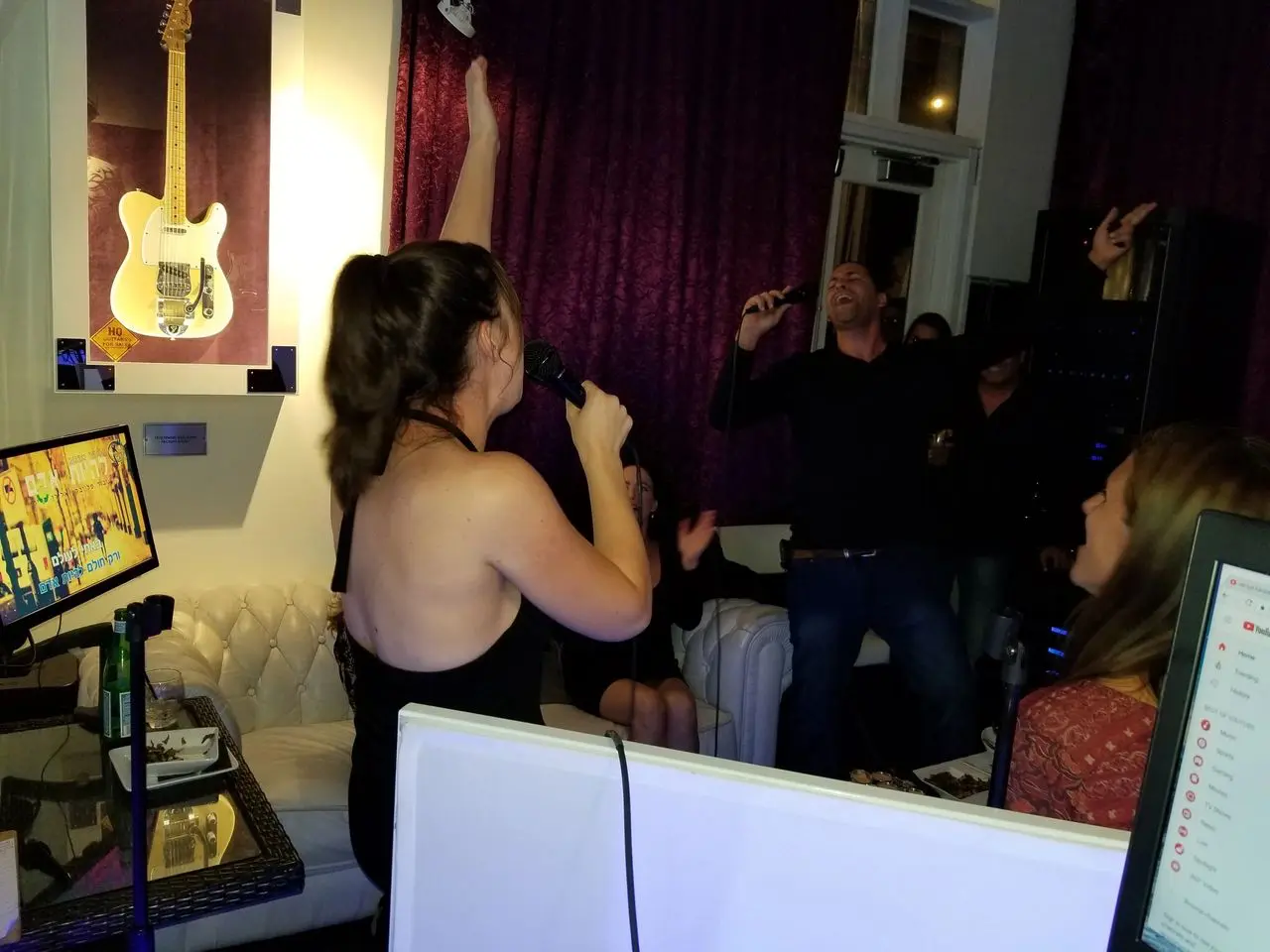 Jewish Karaoke - Israel People singing and cheering in a karaoke room with microphones, a guitar displayed on the wall, and screens showing lyrics.