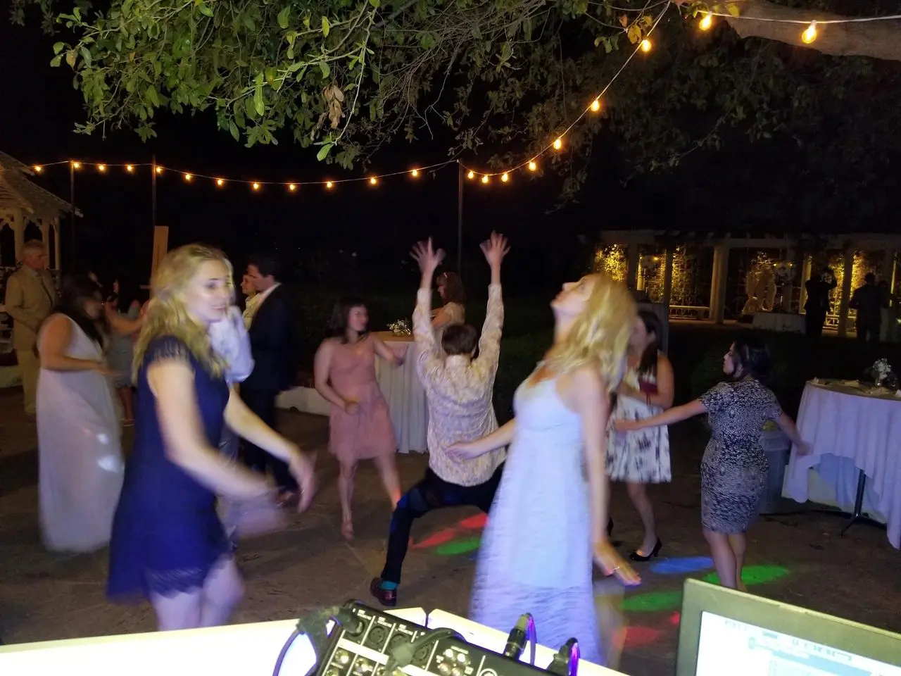 karaoke dj, los angeles dj A group of people dance energetically on an outdoor patio at night, surrounded by string lights, with tables and trees in the background.