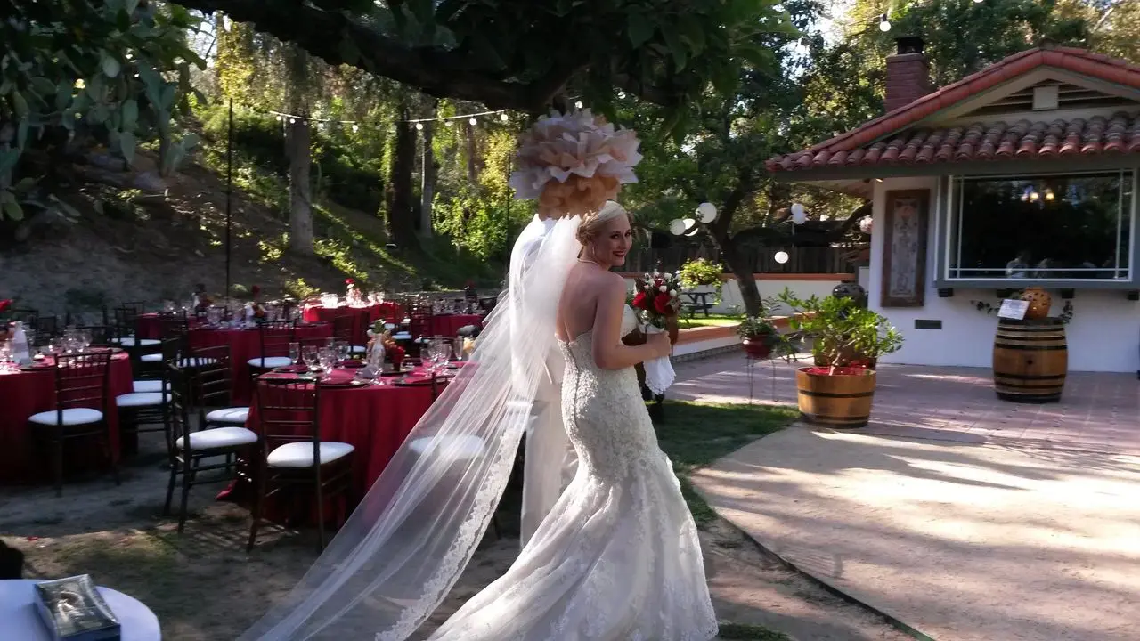 Mr Dj Services - MC Dj A bride in a white gown and veil holds a bouquet and smiles back while walking outdoors at a decorated wedding venue with round tables and red tablecloths.