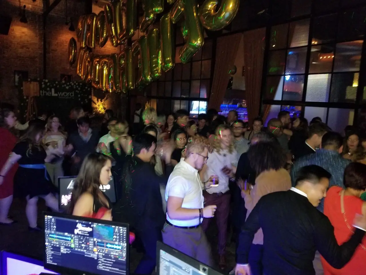 Wrap Party The L Word A large group of people are dancing and socializing in a dimly lit venue with gold balloons and DJ equipment visible in the foreground.