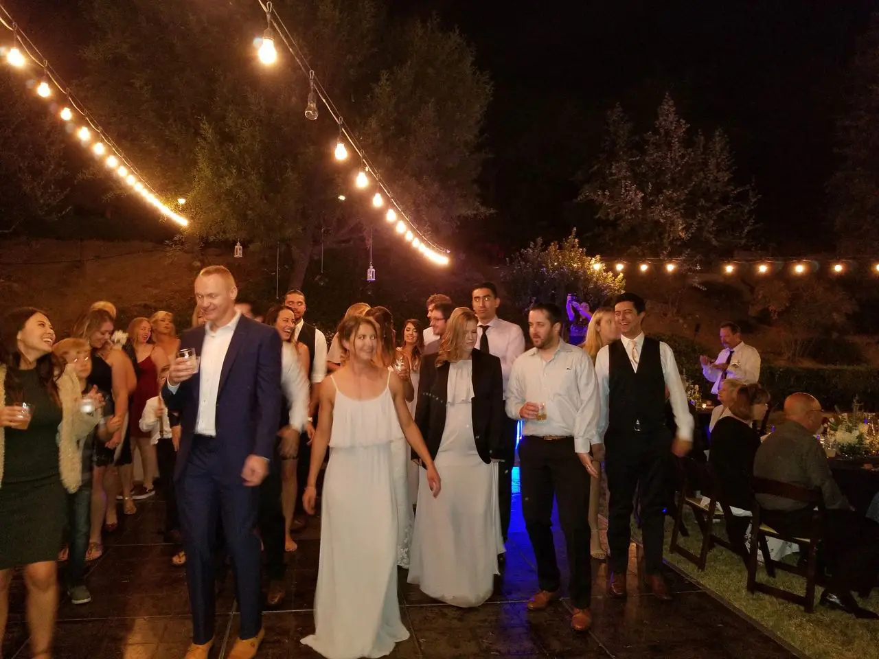 djs for a wedding | wedding dj service A group of people in formal attire walk together on a lit outdoor floor at night, with string lights overhead and trees in the background.