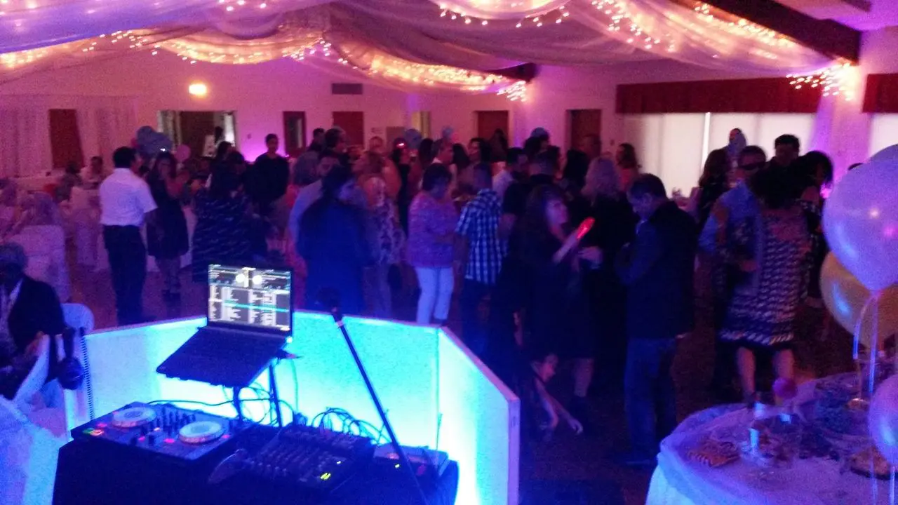 Mr Dj Services in Los Angeles A large group of people are dancing in a decorated banquet hall with string lights, viewed from behind a DJ booth with turntables and a laptop.