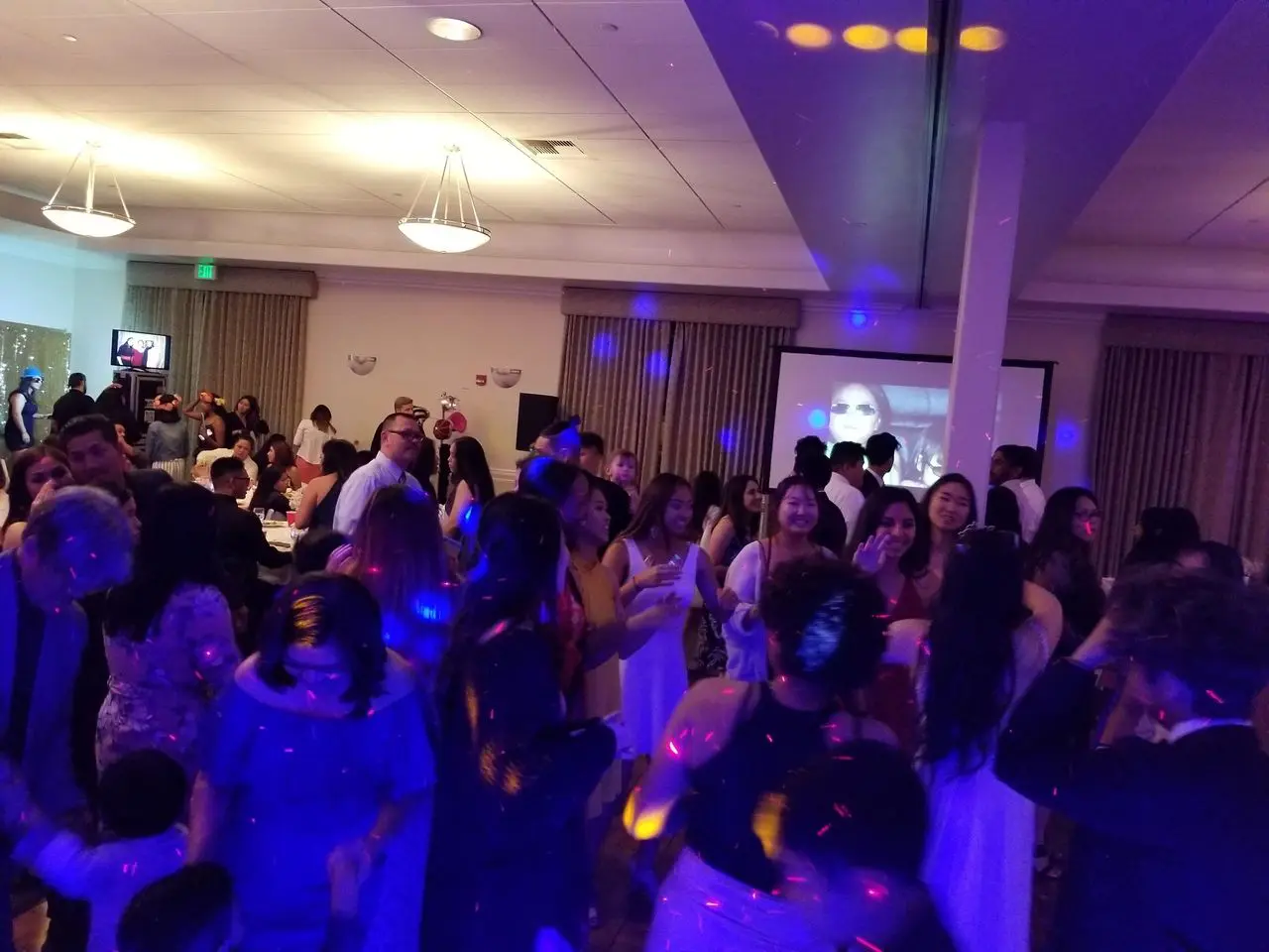 Dj Services - Grad Party and family A large group of people are dancing and socializing in a banquet hall with colorful lights and a projection screen in the background.