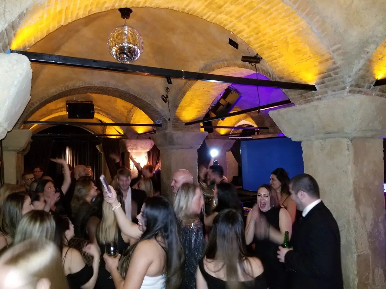 The Hollywood Roosevelt A group of people dressed in formal attire are dancing and socializing at a lively party in a stone-arched room with yellow lighting and a disco ball.