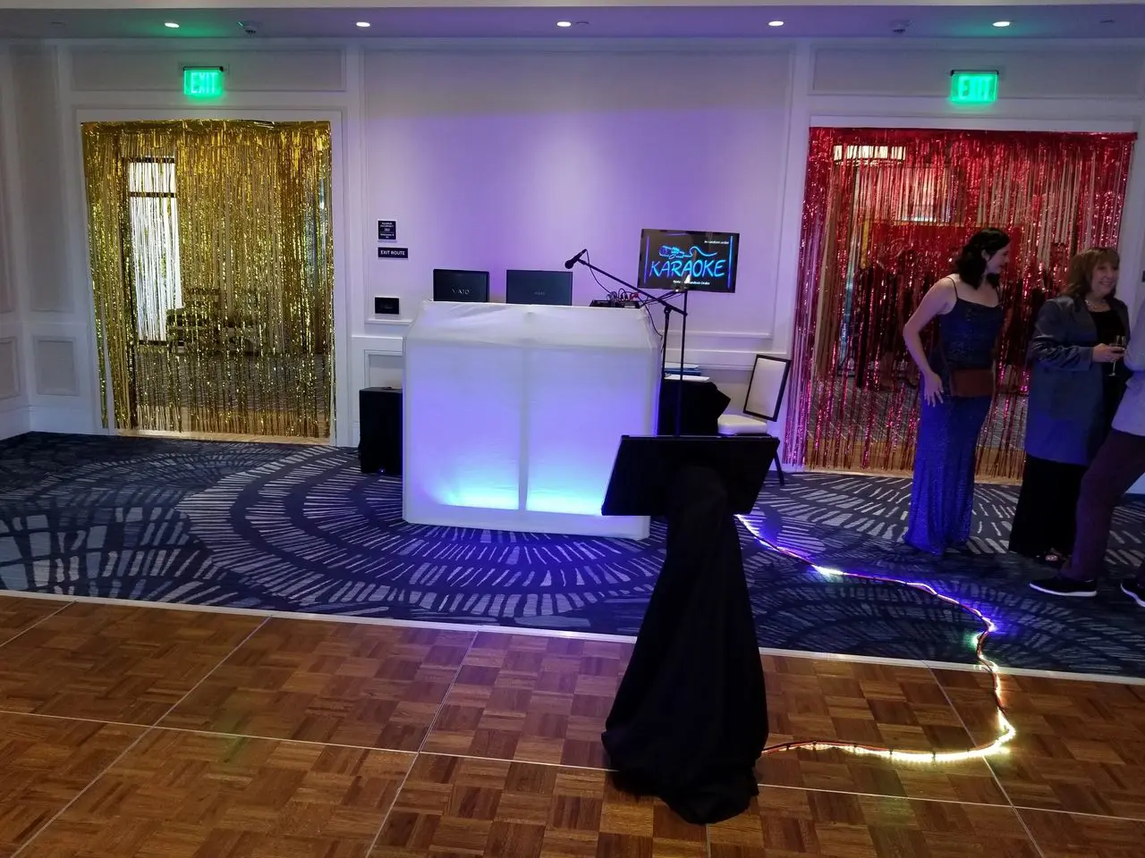 Karaoke Dj Photobooth A karaoke setup with a lit DJ table, screen, and microphone stand on a dance floor; gold and red metallic streamers hang over two doorways; a few people stand nearby.