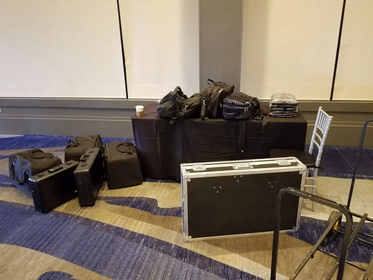 rs=w_1280 (30) A table and floor hold several black equipment cases, bags, folded cloths, and a cup; a folding chair is nearby on a blue-patterned carpet.