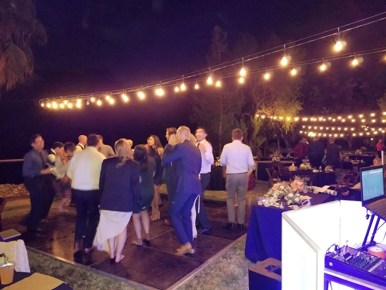 Best wedding dj | los angeles wedding dj A group of people are dancing on an outdoor dance floor at night under string lights, with tables and a DJ booth nearby.
