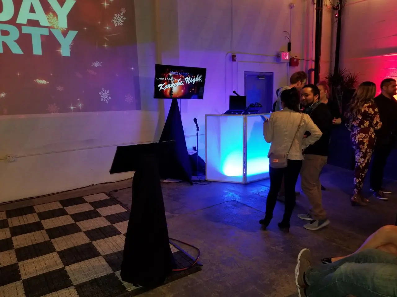 Mr Dj Services A group of people stands near a brightly lit DJ booth and karaoke setup in an indoor event space with party-themed decorations.