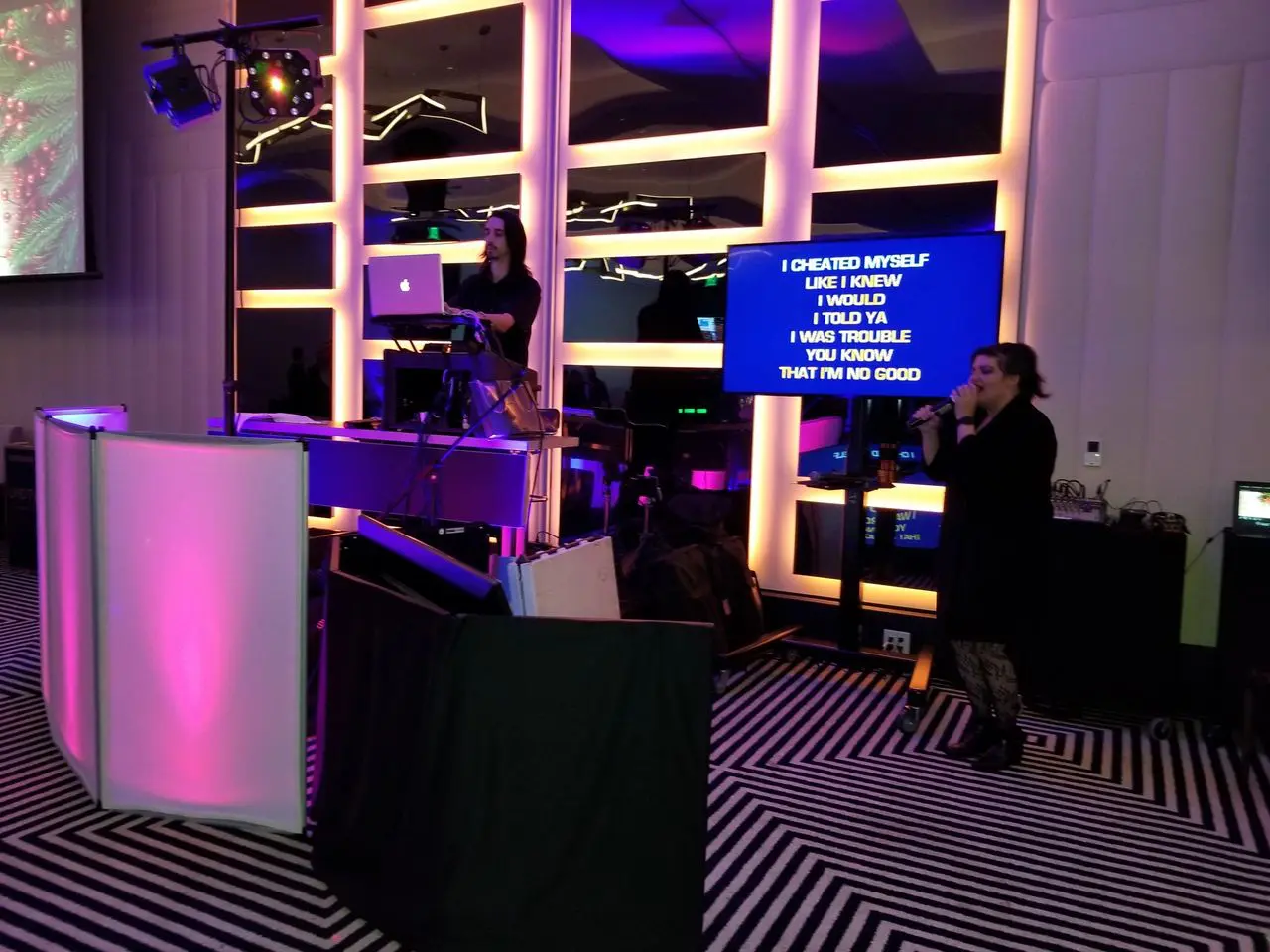Mr Dj Services A woman sings karaoke while another operates a laptop at a DJ booth; song lyrics are displayed on a screen beside her in a room with colorful lighting.