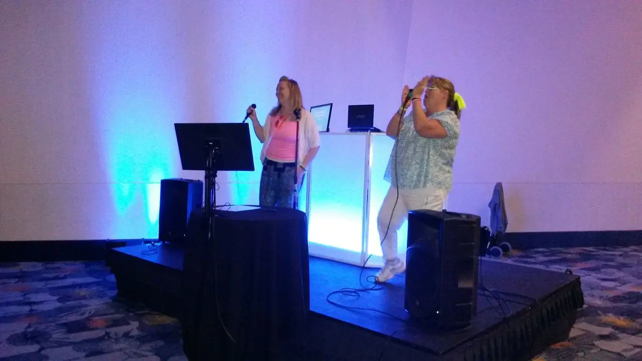 dj and karaoke Two women stand on a small stage singing into microphones, with speakers and a screen in front of them and blue lighting behind them.