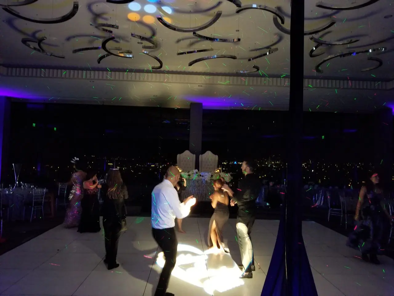 New Year Wedding Dj People are dancing on a dimly lit dance floor with colorful lights and cityscape visible through large windows in the background.