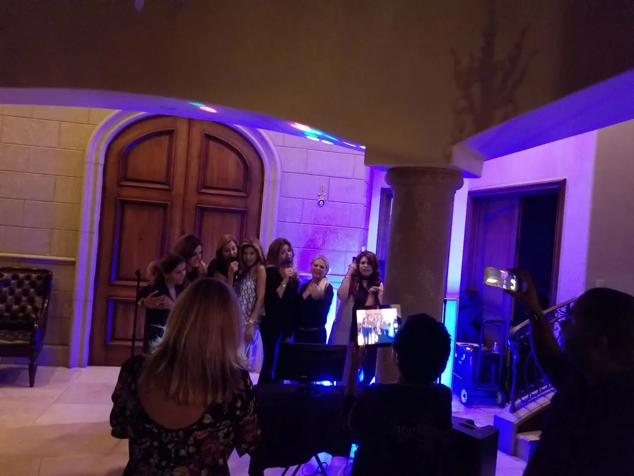 karaoke djs | dj service A group of women stand together singing or speaking into microphones in a warmly lit room, while others watch and take photos or videos.