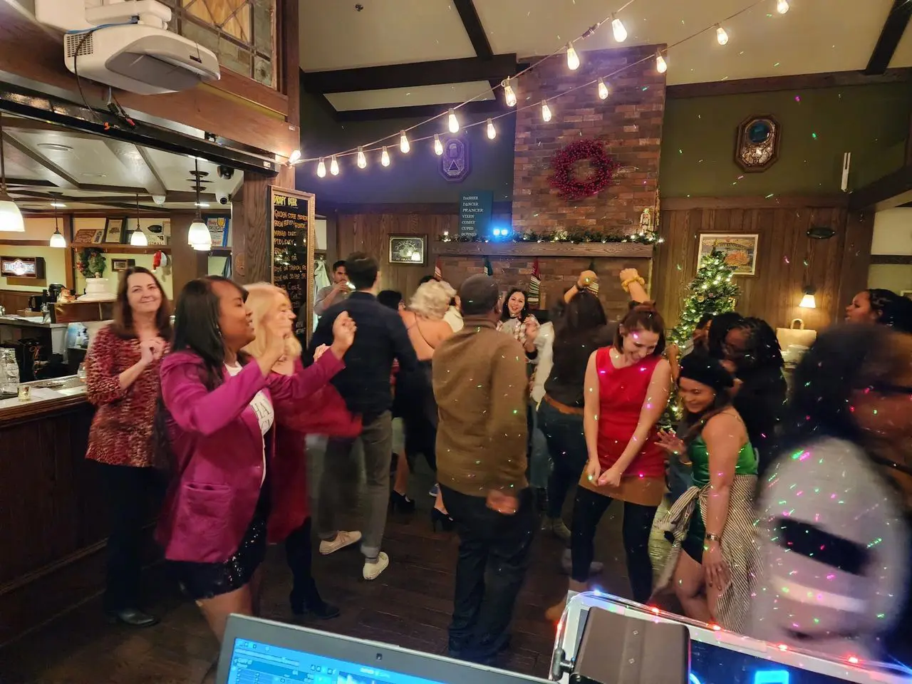 Mr Dj Services A group of people dance and socialize in a warmly lit indoor venue decorated with string lights and holiday ornaments.