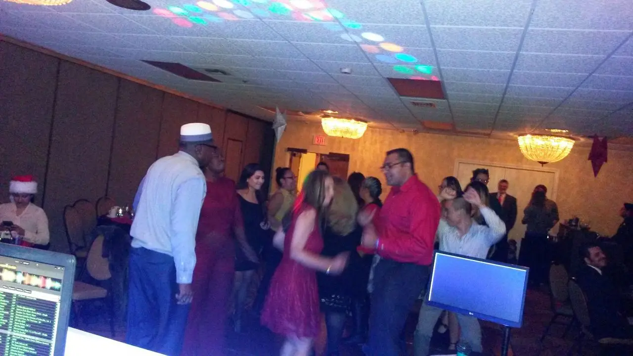 Holiday Dance and Karaoke Dj A group of people are dancing in a dimly lit event room with chandeliers, while others sit at tables. A DJ booth and computer screens are visible in the foreground.