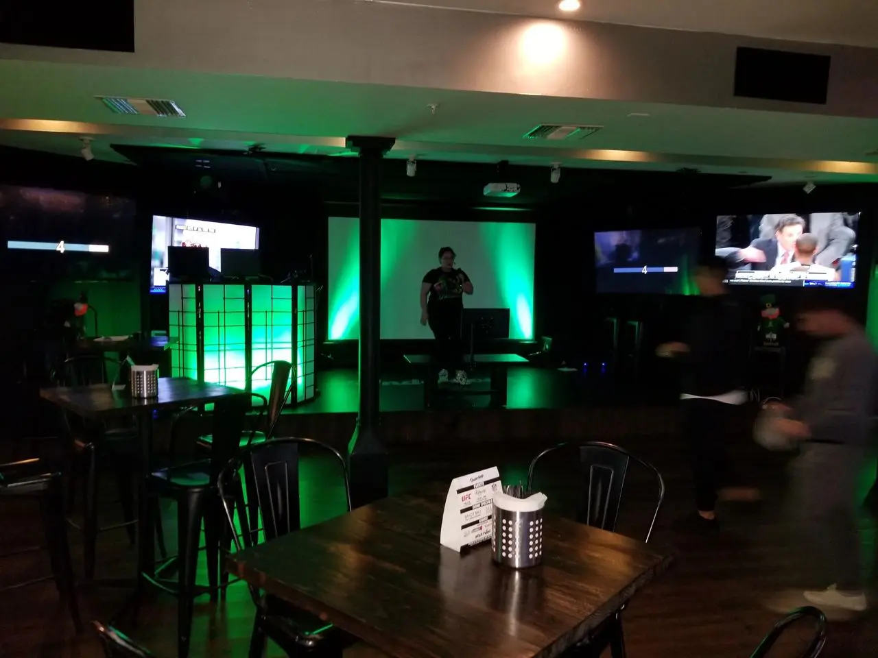 Pre Pandemic Mar 2020 Dimly lit bar with empty tables and chairs, green-lit stage in the center, one person on stage, several TV screens on surrounding walls, and a few people moving around.