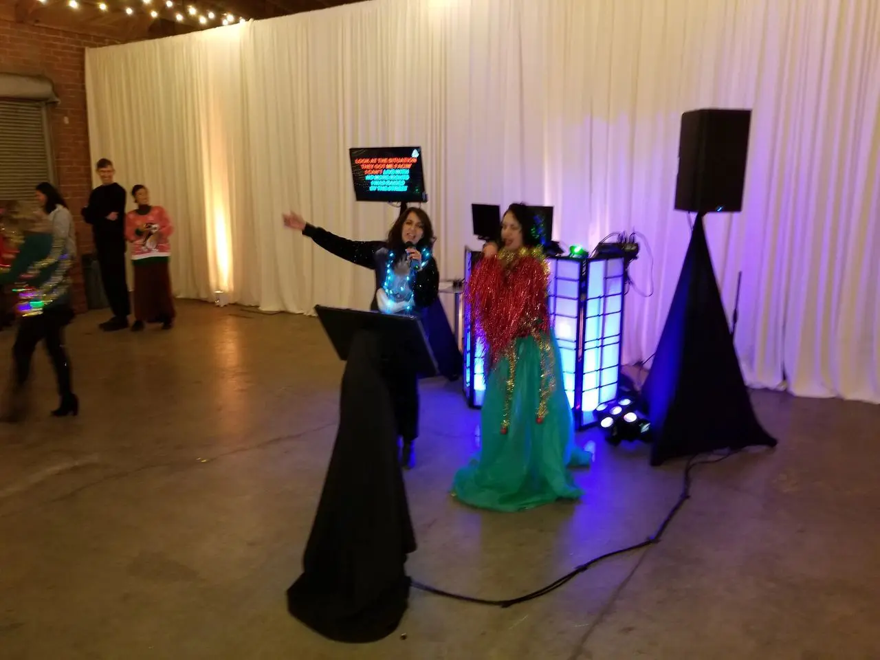 Corporate Party Xmas Two people sing karaoke on microphones in a decorated indoor space, with a karaoke screen, speakers, and colored lights behind them. Other people stand and watch.