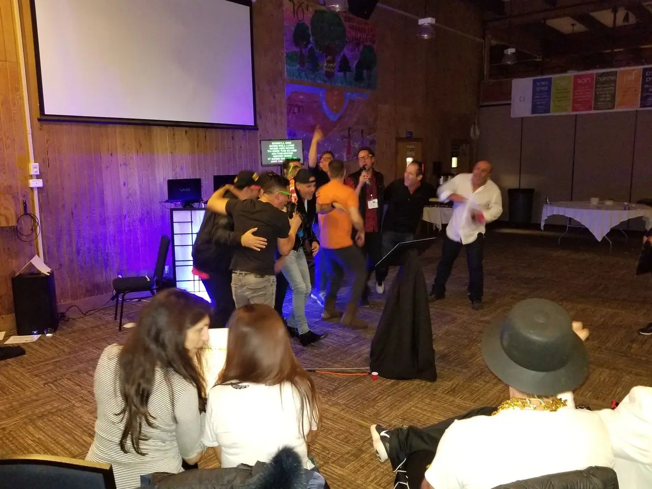 karaoke dj, los angeles dj A group of people gather on a stage under a projector screen, some hugging and celebrating, while others sit and watch in a warmly lit room with wood-paneled walls.