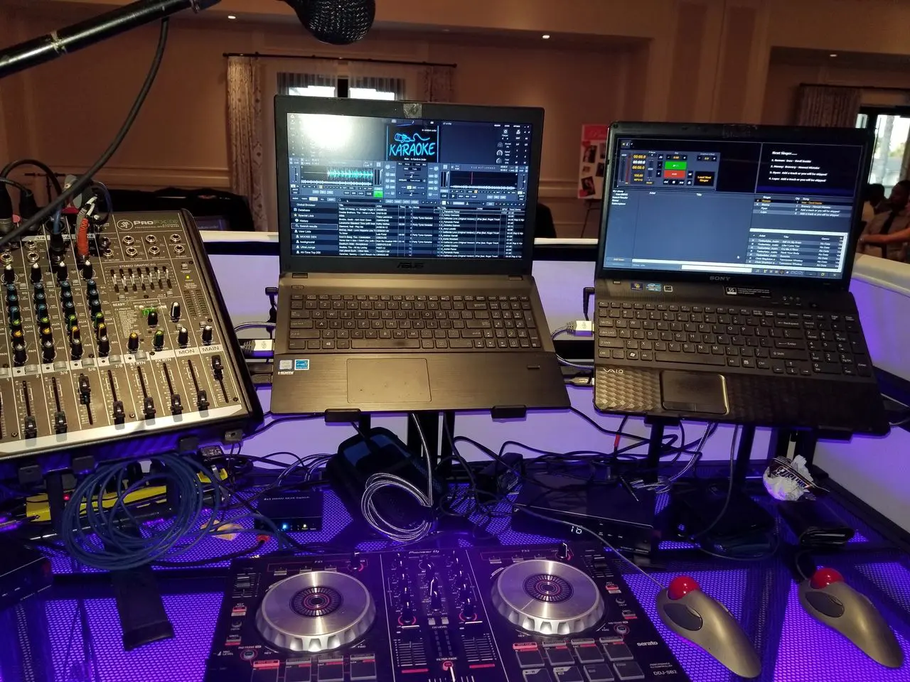 Mr Dj Services DJ and karaoke setup with a mixer, two laptops displaying DJ and karaoke software, a microphone, cables, and a DJ controller on a lit table.