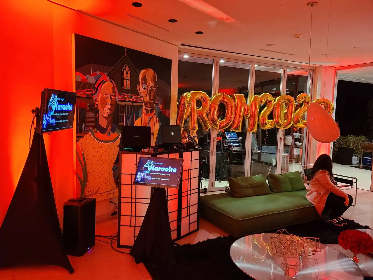 AirBnB rental A living room decorated with "PROM 2023" gold balloons, a karaoke setup, colorful lighting, and a mural; a person sits on a green couch.